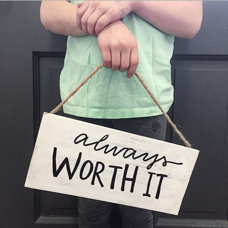 Close up view of a person standing in front of a door, holding a sign that says, "Always worth it."