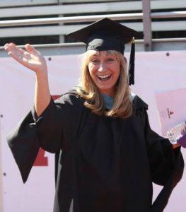 Blonde woman in black graduation cap and gown waving to camera.