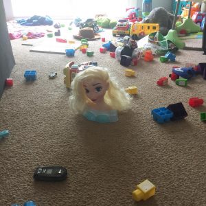 Kids toys including an Elsa barbie head, blocks, toy cell phone, and trucks laid out all over the living room floor.