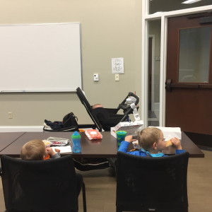 Baby in a stroller, two young blonde toddlers in college classroom chairs eating dinner.