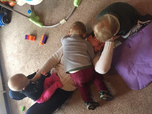 Woman laying on carpeted floor with three little kids piling on top of her. A baby girl at her knees, toddler boy on her chest, and another toddler boy on her face.