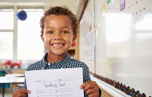 young boy smiling while holding up spelling test